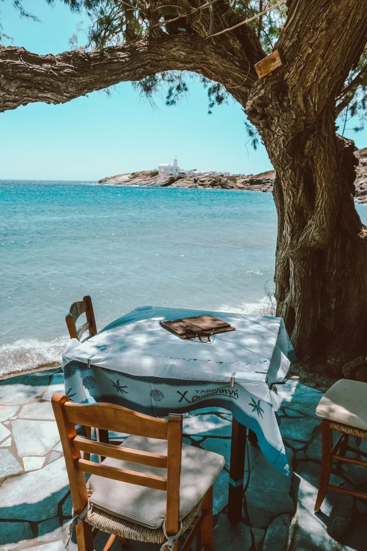 A taverna table under a tamarisk tree at the water's edge, with Chrysopigi monastery beyond
